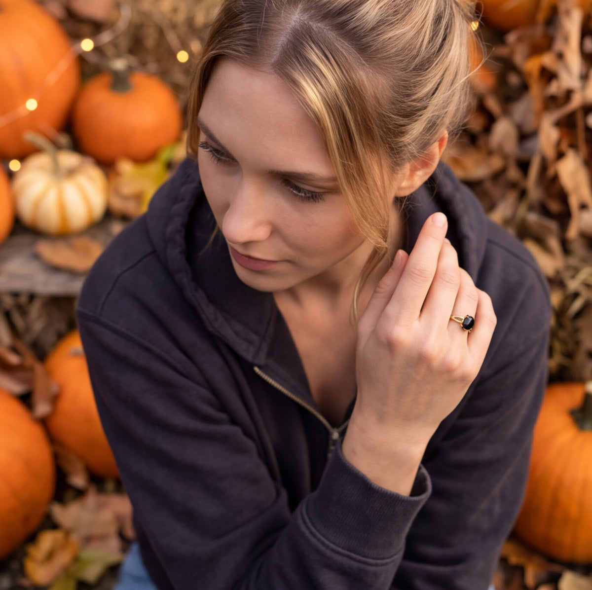 A woman in a dark navy zip-up hoodie poses outdoors among pumpkins and autumn leaves, wearing a black onyx ring with a clear central gemstone on her finger.