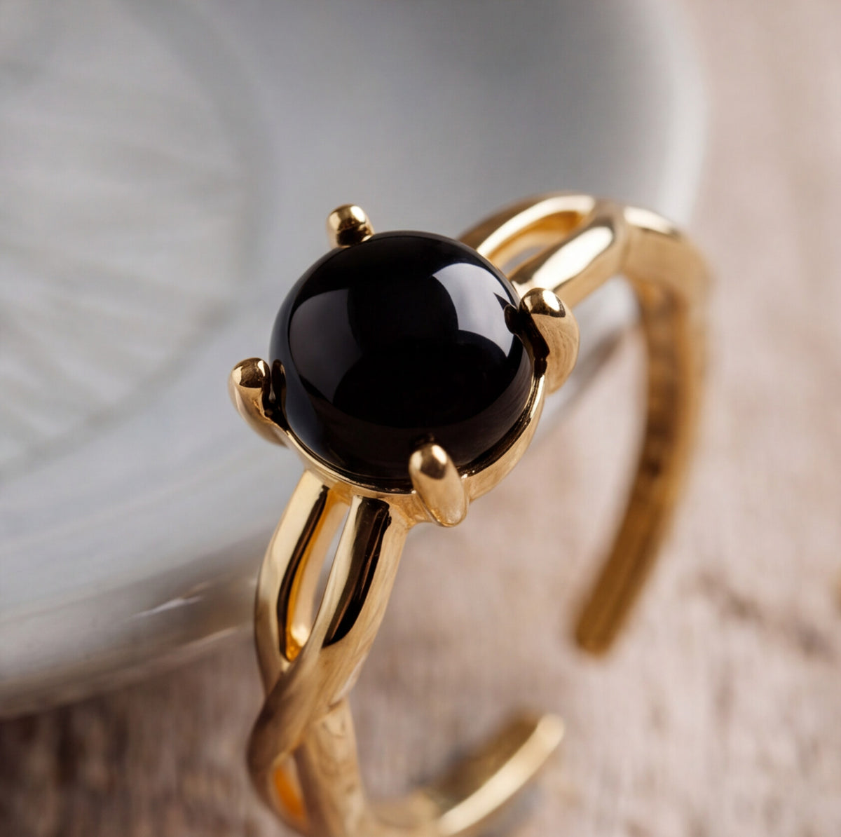 A gold-toned open cuff ring featuring a large, smooth, polished black oval onyx gemstone in a prong setting, resting on a light textured surface.