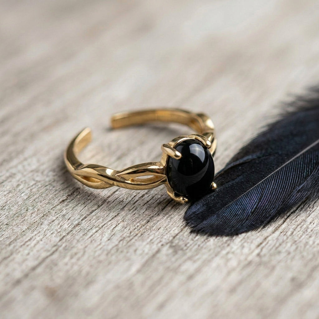 A gold-toned twisted band ring with a smooth, polished black oval onyx gemstone, resting on a light wood surface next to a dark feather.