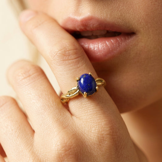 A close-up lifestyle shot of a gold ring with an oval blue lapis lazuli stone worn on a finger, with a person's lips and face visible in the background.