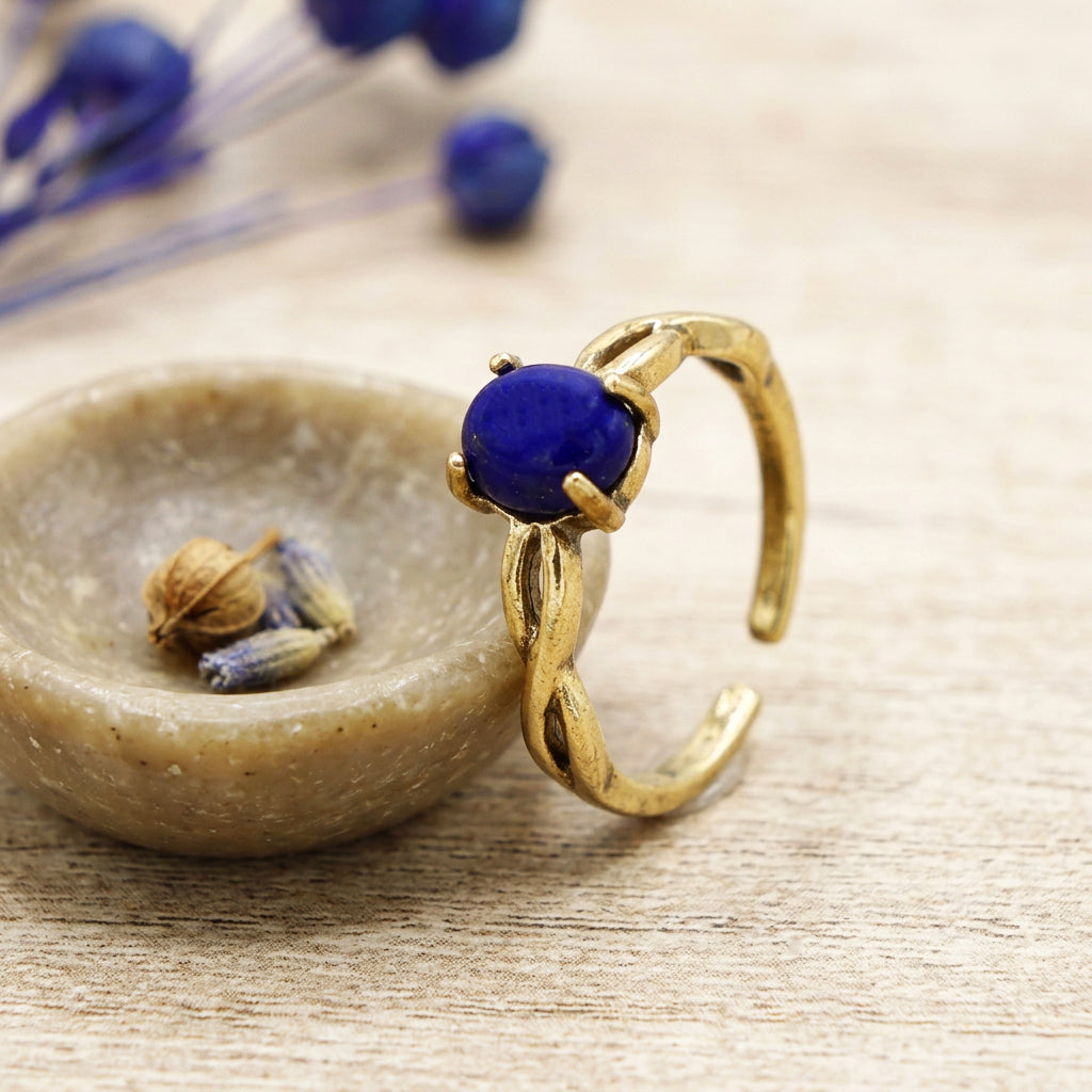 A gold-toned open ring with a round deep blue lapis lazuli gemstone, displayed on a textured beige stone dish with dried lavender on a light wood surface.
