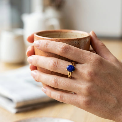 A close-up lifestyle shot of a person's hands holding a speckled brown ceramic mug, wearing a ring with a deep blue oval lapis lazuligemstone on one finger.
