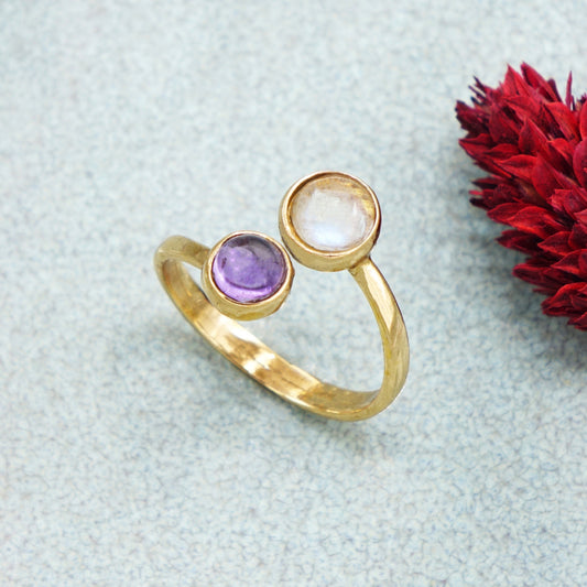 Gold ring with two gemstones on a light blue textured surface with a red flower.