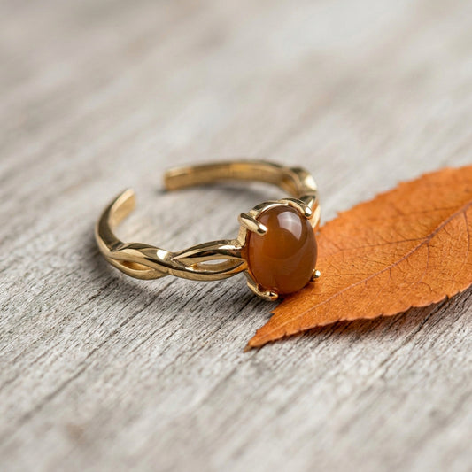 A gold-toned open ring with a smooth oval amber-brown agate gemstone, photographed on a light wood surface next to an orange leaf.