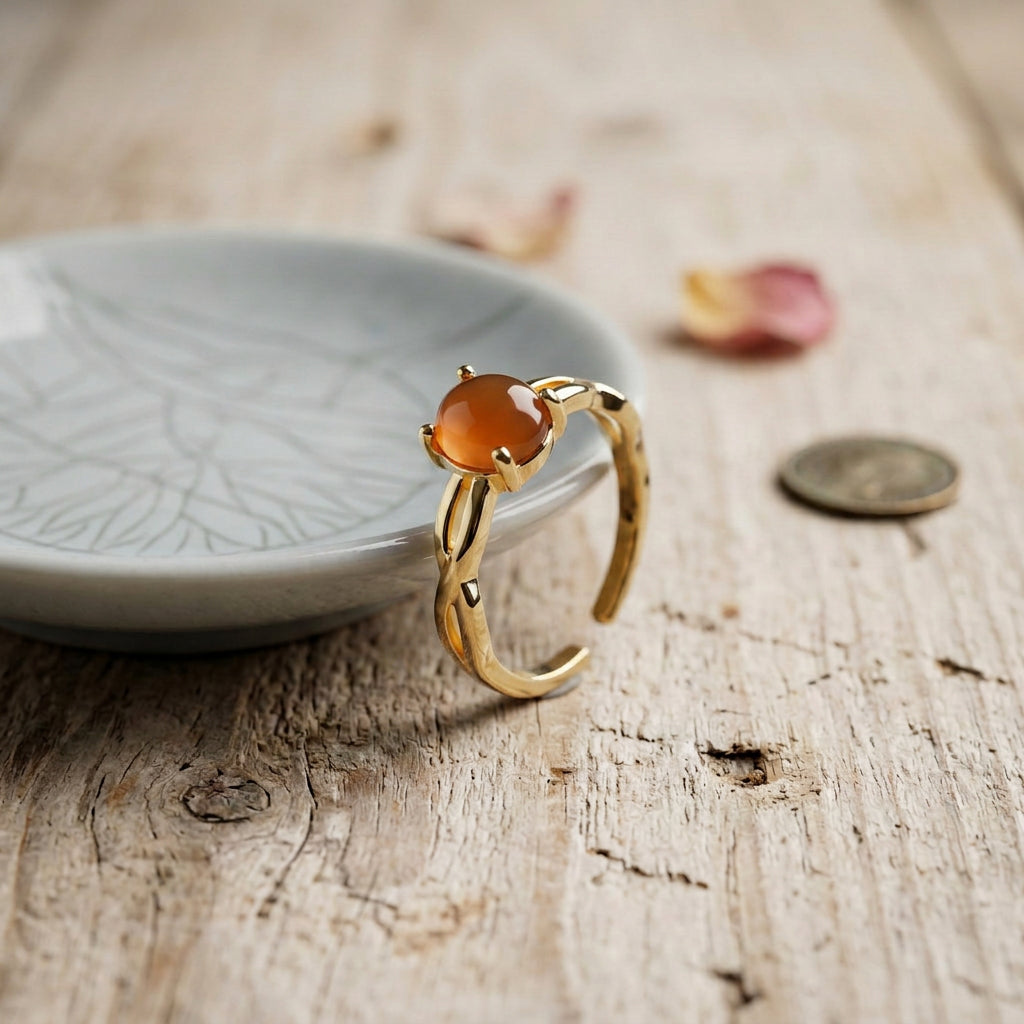 A gold ring with a round amber-orange cabochon gemstone, displayed on a rustic wooden surface next to a small ceramic dish.