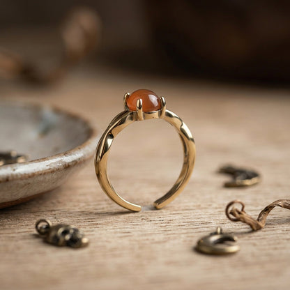 A gold-toned adjustable ring with a smooth, oval amber-orange agate gemstone, displayed on a rustic wooden surface with jewelry findings nearby.