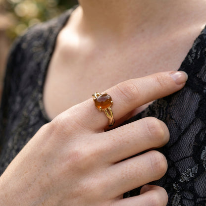 A gold ring with a heart-shaped amber cabochon gemstone worn on a person's finger, set against a black lace top and a soft, blurred outdoor background.