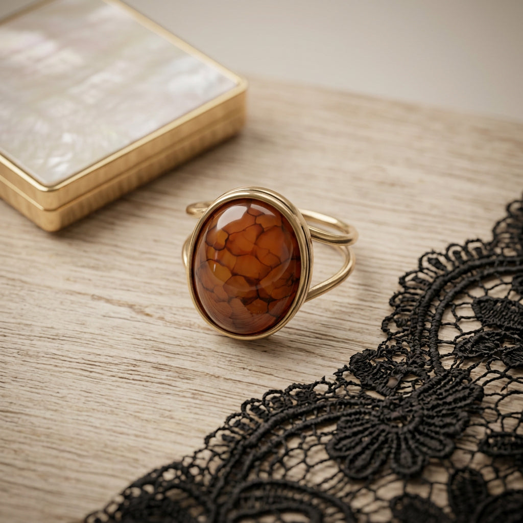 A gold ring with a large oval agate cabochon stone, styled on a light wood surface next to a black lace trim and a gold-toned box.