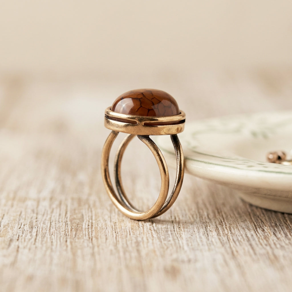 A gold-toned double-band ring with a polished dragon vein oval agate cabochon stone, displayed on a light wood surface next to a ceramic dish.