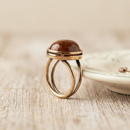 A gold-toned double-band ring with a polished dragon vein oval agate cabochon stone, displayed on a light wood surface next to a ceramic dish.