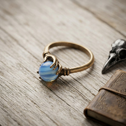 A bronze ring featuring a carved hand holding a round, moonstone gemstone, displayed on a rustic wooden surface next to a bird skull and an antique book.