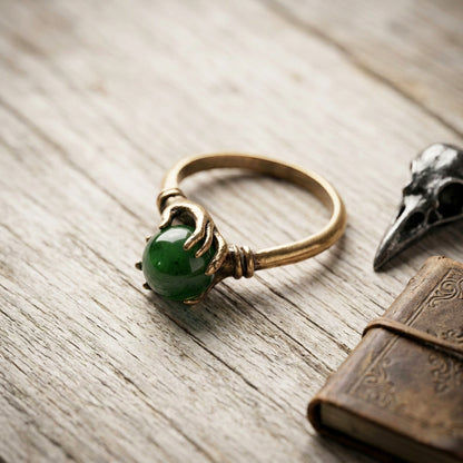 A gothic-style ring featuring a polished green jade stone held by two detailed gold fingers, styled on a rustic wooden surface next to a bird skull and an antique book.