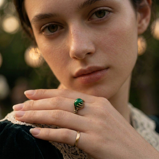 A close-up lifestyle shot of a woman wearing a bronze ring with a green jade stone on her middle finger, with a second thin gold band on her ring finger, set against a soft, blurred background.