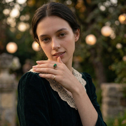 A woman in a dark green velvet dress with a cream lace collar wears a green jade gemstone ring on one finger, posed outdoors against a soft-focus background of trees and warm lights.
