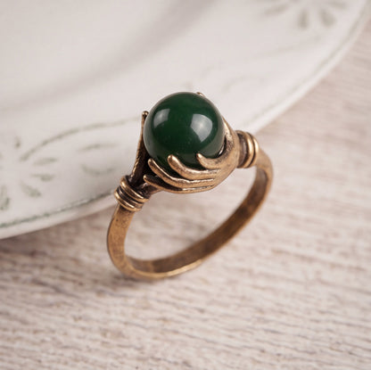 A vintage-style ring featuring a round, polished dark green jade stone held by two antique bronze hands, resting on a light wood surface next to a white ceramic plate.