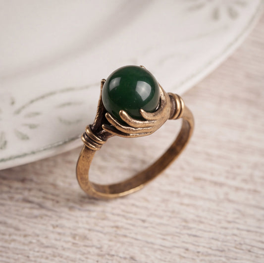 A vintage-style ring featuring a round, polished dark green jade stone held by two antique bronze hands, resting on a light wood surface next to a white ceramic plate.
