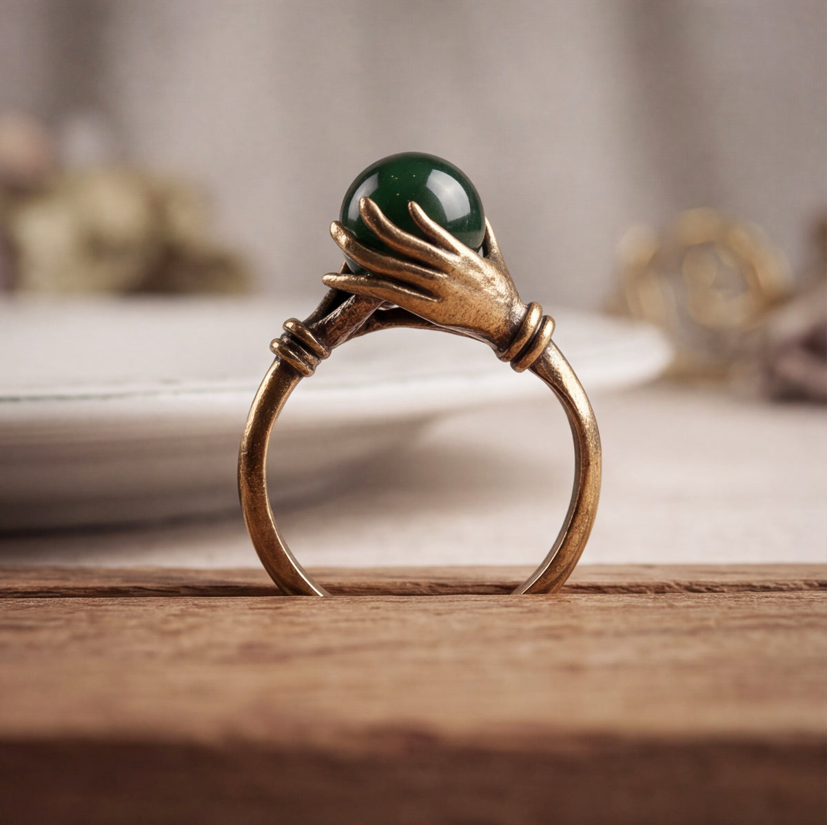 A gold ring featuring two sculpted hands holding a round green jade stone, displayed on a wooden surface with a softly blurred background.