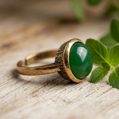 A gold-toned adjustable ring with a large, polished green jade gemstone, resting on a rustic wooden surface next to green leaves.