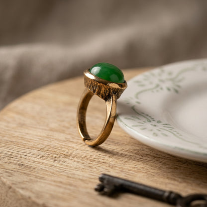 A gold ring with a smooth green oval jade stone, displayed on a wooden surface next to a white plate and a vintage key.