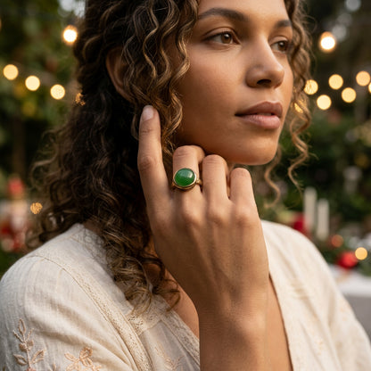 A close-up lifestyle shot of a woman with curly brown hair wearing a gold ring with a green oval stone, set against a softly blurred background with warm bokeh lights.