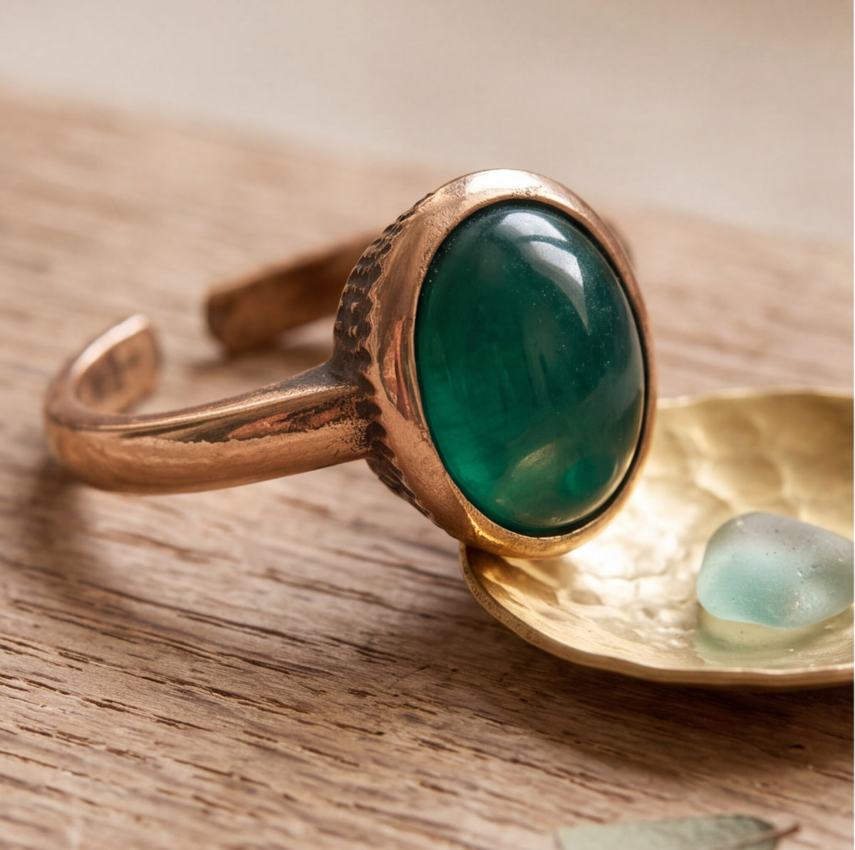 A close-up of a gold-toned adjustable ring with a large oval green cabochon stone, resting on a textured gold dish over a wooden surface.
