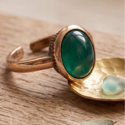 A close-up of a gold-toned adjustable ring with a large oval green cabochon stone, resting on a textured gold dish over a wooden surface.