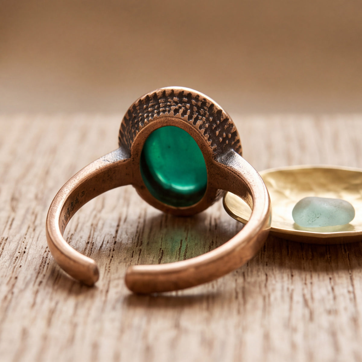 A close-up of a handmade copper-toned ring with a textured circular setting and a smooth green oval stone, resting on a wooden surface next to a small gold dish with a light blue stone.