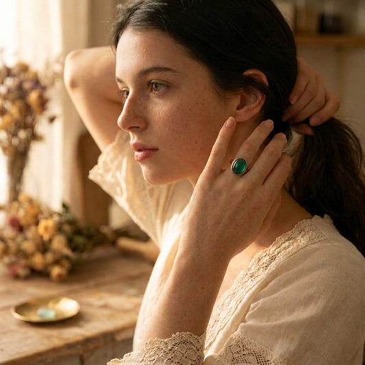 A woman in a cream lace blouse poses in a warm, softly lit room, wearing a prominent ring with a large oval green stone on her finger.