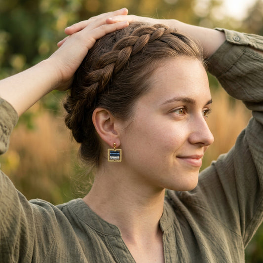 A woman with a braided brown ponytail wearing square gold-tone earrings with blue and white striped details, posing outdoors with her hands in her hair.