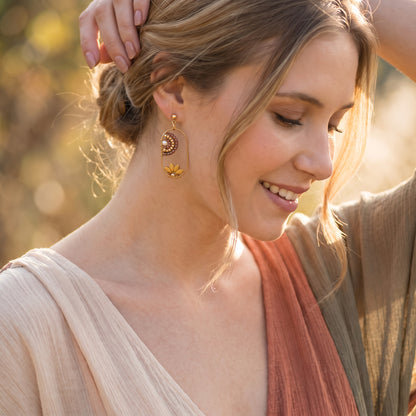 A woman wearing gold crescent moon and flower dangle earrings, shown in a warm outdoor setting with soft bokeh background.
