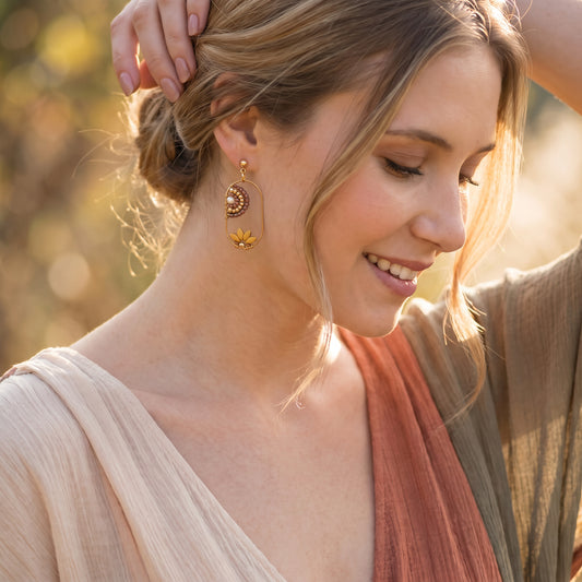 A woman wearing gold crescent moon and flower dangle earrings, shown in a warm outdoor setting with soft bokeh background.