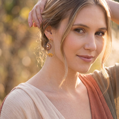 A woman wearing a pair of gold-toned spiral drop earrings with brown detailing, photographed outdoors with a warm, blurred natural background.
