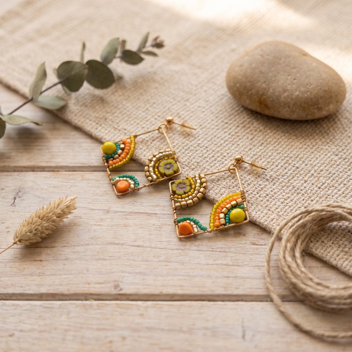 A pair of gold-toned beaded dangle earrings with colorful geometric and rainbow-like patterns in green, orange, yellow, and purple, displayed on a rustic wooden surface with burlap, a stone, eucalyptus leaves, and twine.