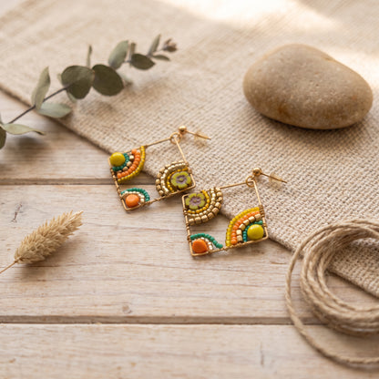A pair of gold-toned beaded dangle earrings with colorful geometric and rainbow-like patterns in green, orange, yellow, and purple, displayed on a rustic wooden surface with burlap, a stone, eucalyptus leaves, and twine.
