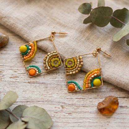 A pair of gold-toned beaded dangle earrings with geometric and floral motifs in orange, yellow, and teal, styled on a rustic wooden surface with a linen cloth and eucalyptus leaves.