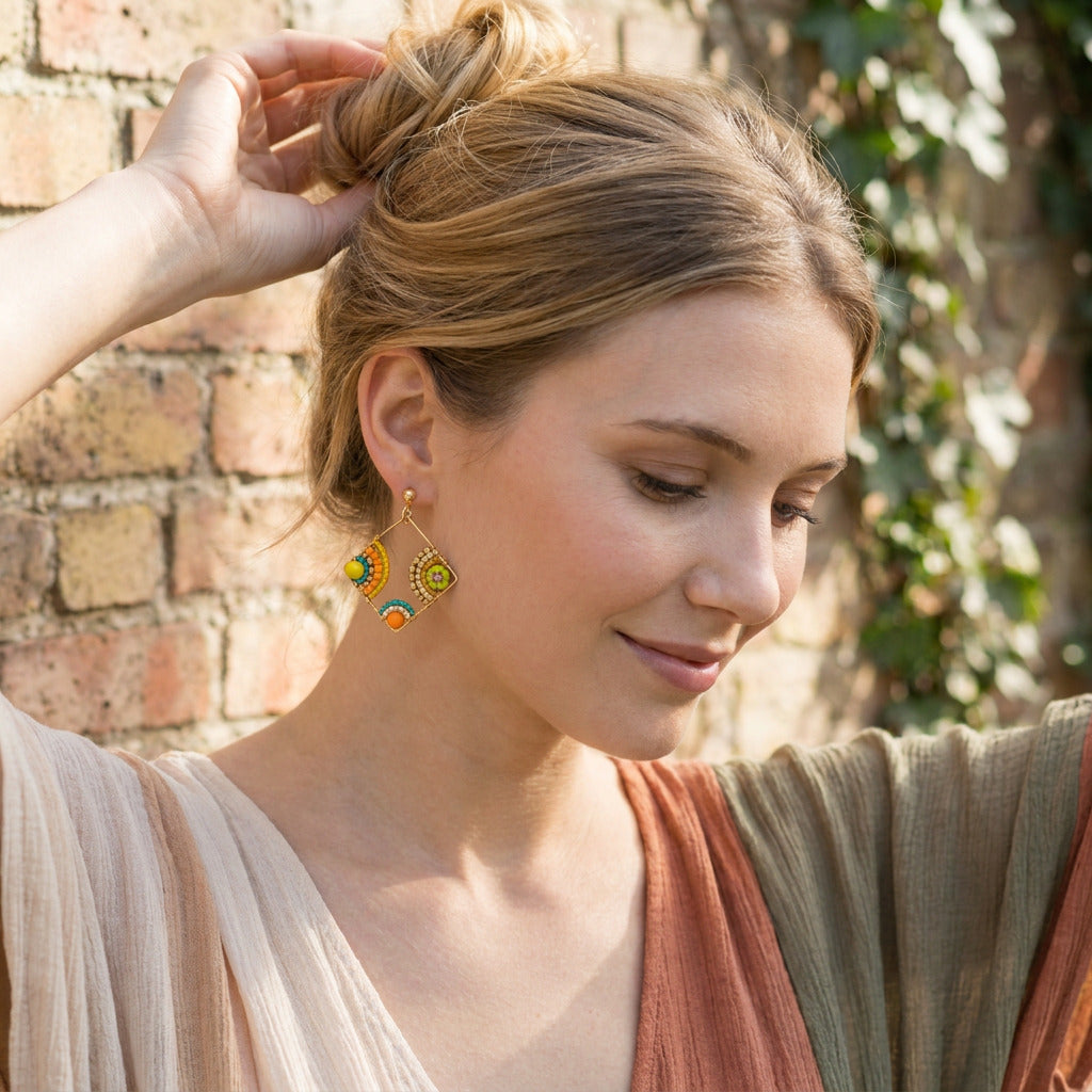 A woman with blonde hair in a bun wearing colorful beaded dangle earrings, posing outdoors against a brick wall and green foliage background.