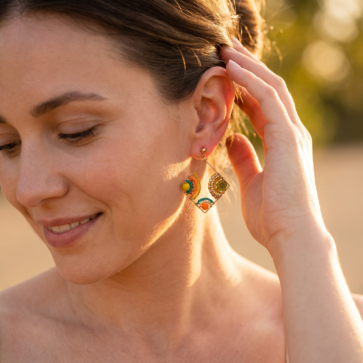 A close-up lifestyle shot of a woman wearing gold-toned geometric dangle earrings with colorful enamel details, set against a warm, blurred outdoor background.