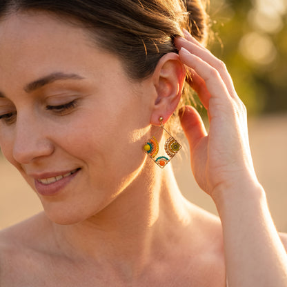 A close-up lifestyle shot of a woman wearing gold-toned geometric dangle earrings with colorful enamel details, set against a warm, blurred outdoor background.