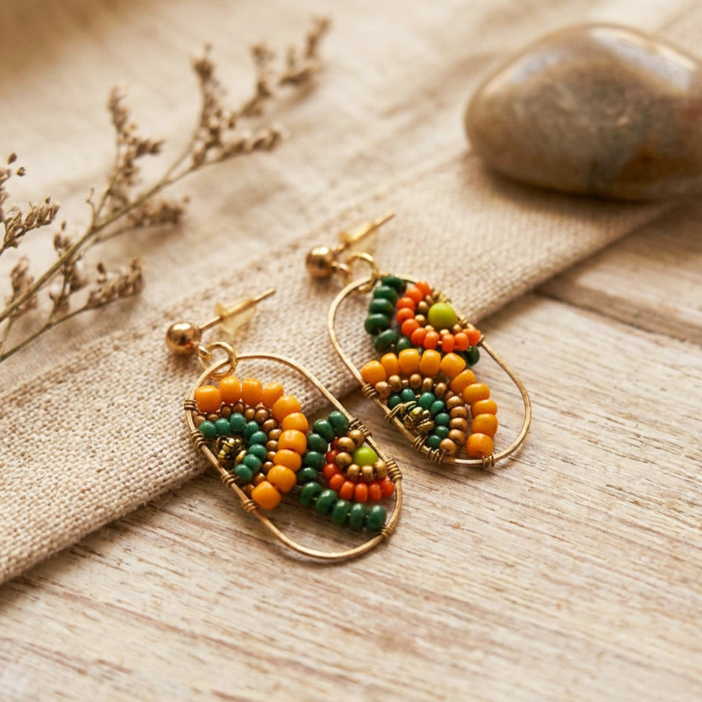 A pair of handmade gold-tone hoop earrings with colorful seed beads in orange, green, and yellow, displayed on a rustic beige fabric and light wood surface.