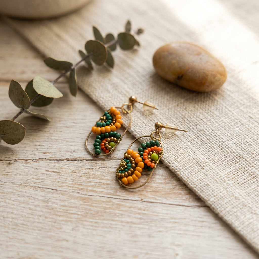 A pair of gold-tone dangle earrings with green, orange, and yellow beaded floral details, displayed on a textured beige fabric over a light wood surface with eucalyptus leaves and a stone.