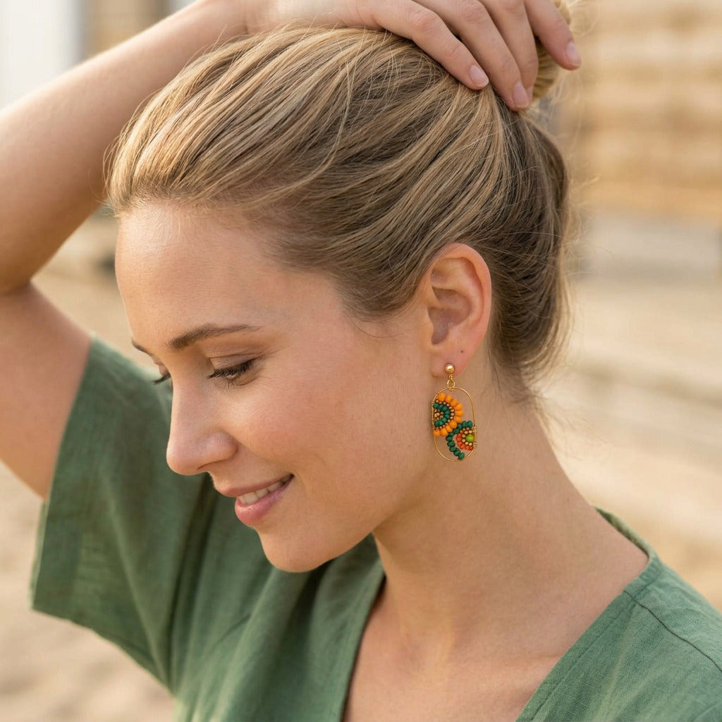 A woman with blonde hair in a bun wearing a pair of handmade beaded earrings shaped like sunflowers, posed in a soft-focus outdoor setting.
