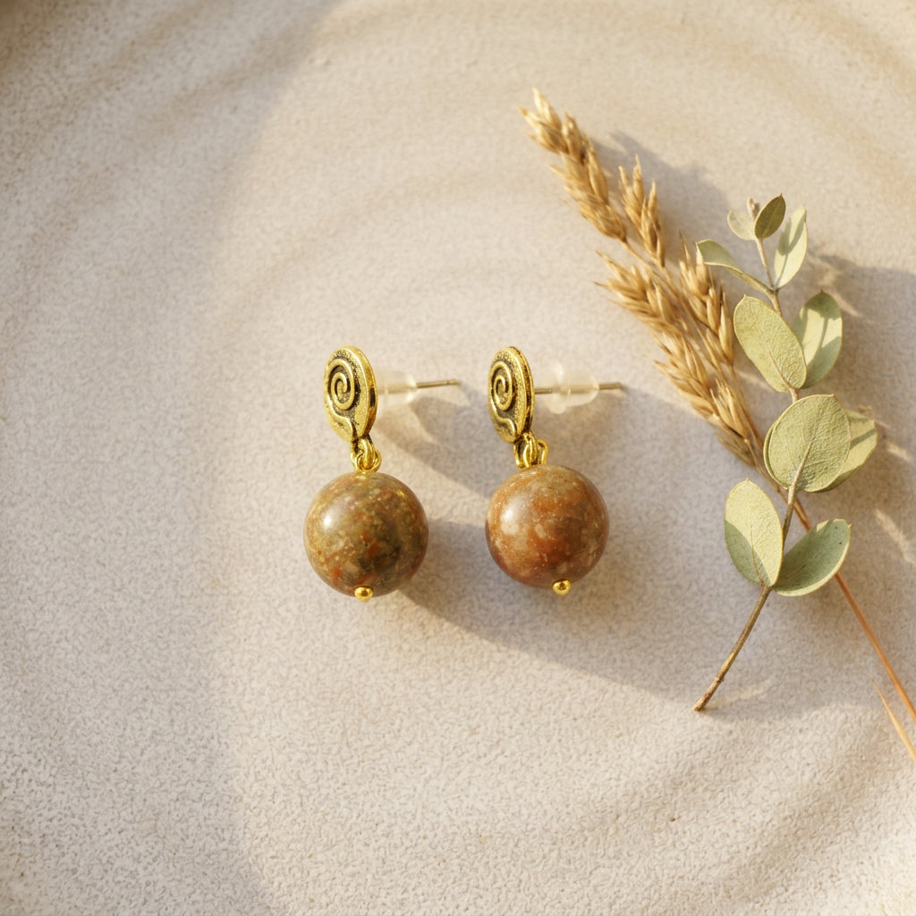 A pair of gold spiral drop earrings with round brown and green jasper stone  pendants, styled on a textured beige surface next to dried wheat and eucalyptus leaves.