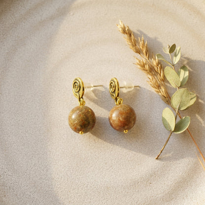A pair of gold spiral drop earrings with round brown and green jasper stone  pendants, styled on a textured beige surface next to dried wheat and eucalyptus leaves.