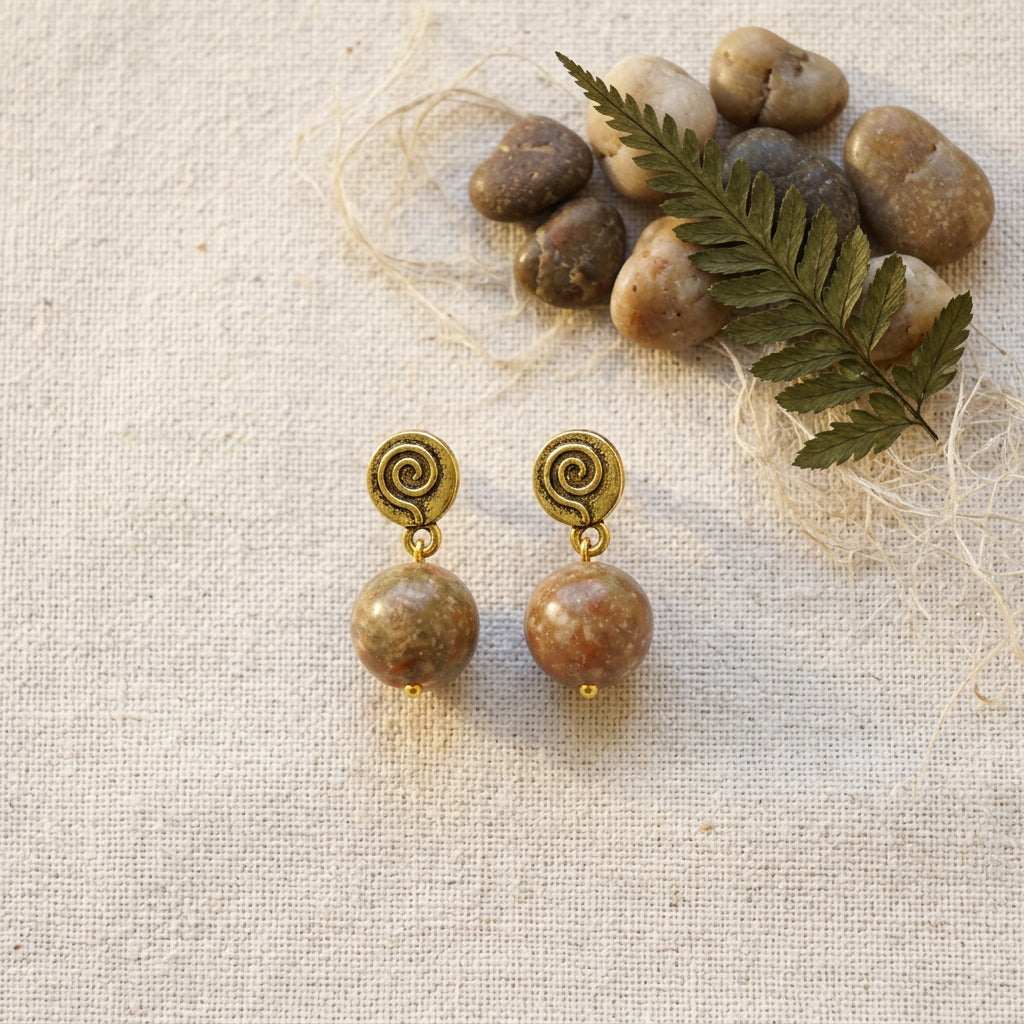A pair of handmade spiral drop earrings with polished jasper stone beads and gold-toned metal caps, displayed on a textured beige linen fabric alongside decorative river stones and a green fern leaf.