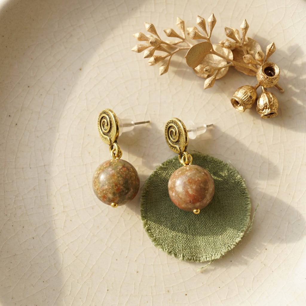 A pair of gold-toned dangle earrings with round brown and green jasper stone beads, displayed on a textured beige ceramic surface next to a gold leaf branch accessory and a green fabric circle.