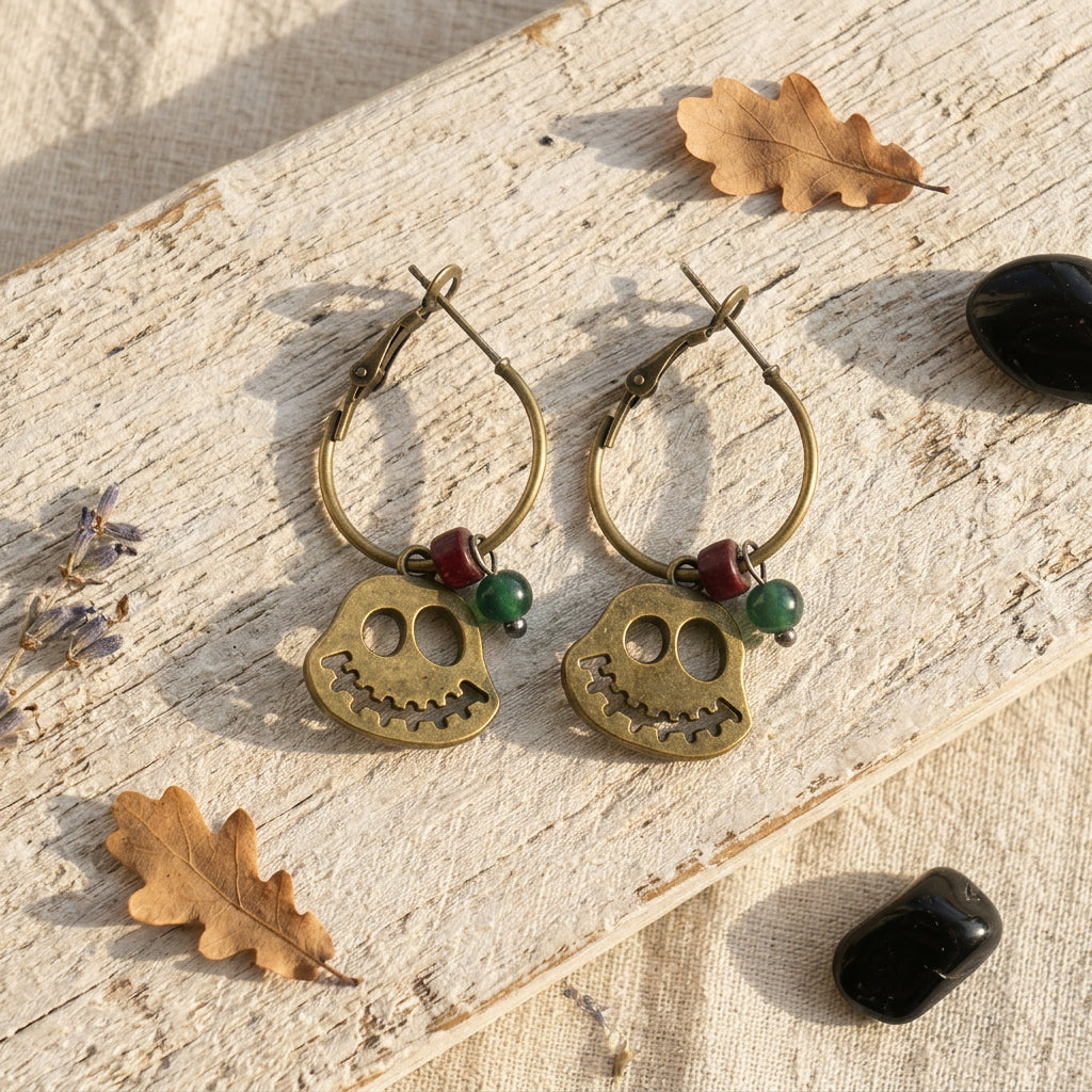 A pair of dangling hoop earrings with ghost charms and small green and red beads, displayed on a weathered wood surface with dried leaves and stones.