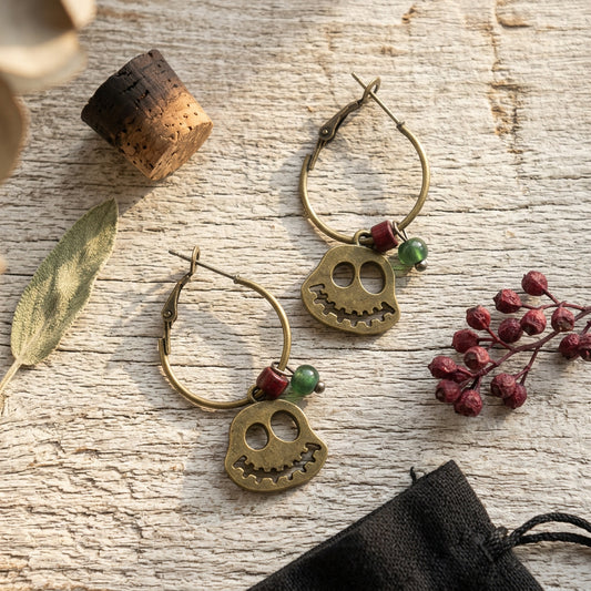 A pair of gold-toned hoop earrings with ghost charms and small red and green beads, displayed on a rustic white wood surface with decorative dried leaves, berries, a cork, and a black drawstring bag.