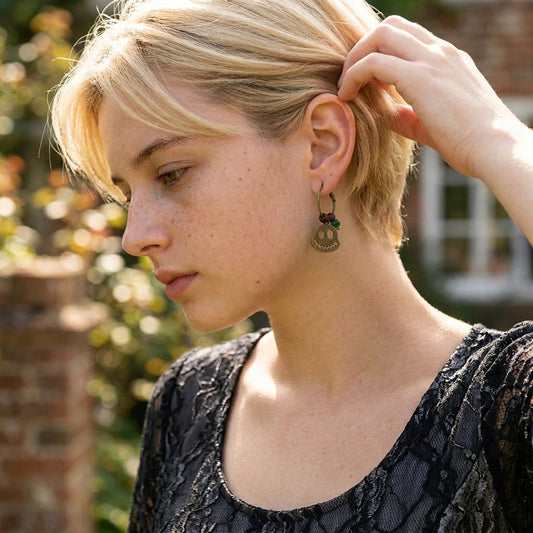 A close-up lifestyle shot of a woman with short blonde hair wearing a small, dangling antique bronze hoop earring with a ghost charm and tiny green bead accents. She is wearing a black lace top, and the background shows a softly blurred brick wall and garden foliage.