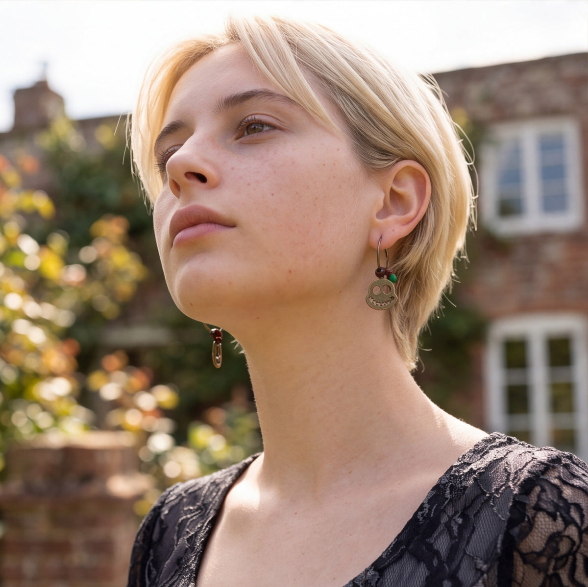A close-up portrait of a woman with short blonde hair wearing a dangling earring with a round silver-toned coin charm and small green and red beads, set against a blurred outdoor background of a brick building and foliage.
