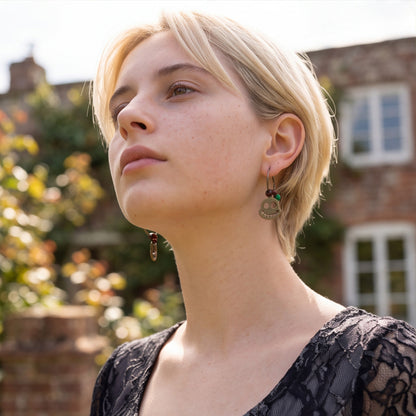 A close-up portrait of a woman with short blonde hair wearing a dangling earring with a round silver-toned coin charm and small green and red beads, set against a blurred outdoor background of a brick building and foliage.